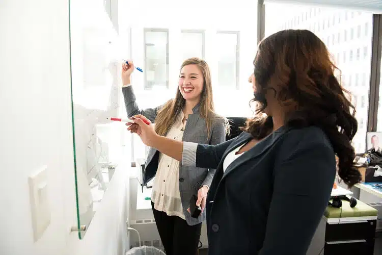 Duas profissionais analisando estratégias em um quadro branco em ambiente corporativo, utilizando ferramentas de gestão para aumentar a produtividade organizacional.